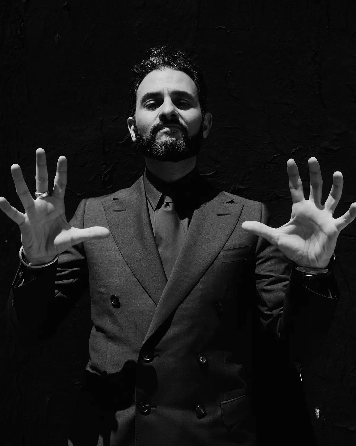 Portrait of a man in a suit standing in front of a black wall with his hands up, showing off his fingers, in black and white.