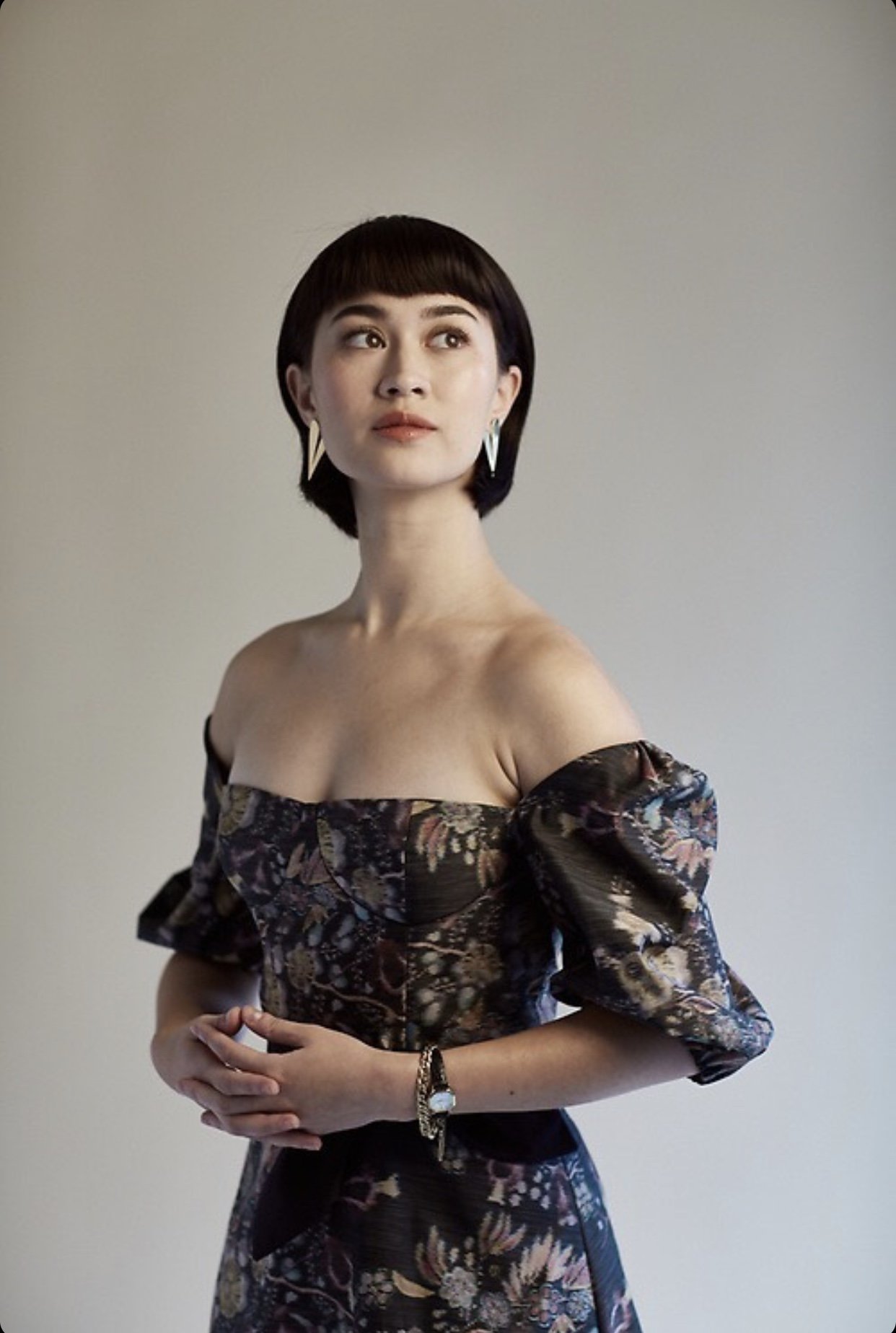 Woman with dark hair and bangs wearing an off-the-shoulder floral dress against a neutral grey studio backdrop.