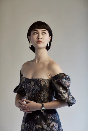 Woman with dark hair and bangs wearing an off-the-shoulder floral dress against a neutral grey studio backdrop.