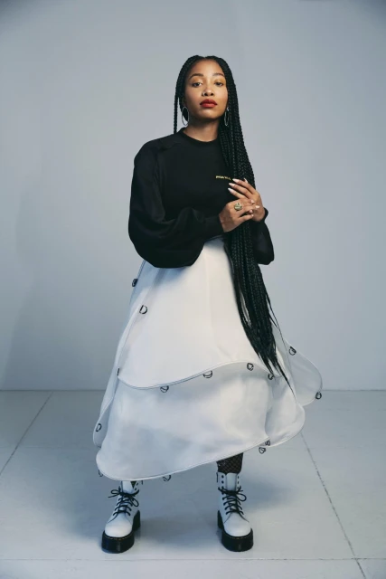 A full shot captures a Black woman in a black top, white skirt, and white boots, posing in a photo studio with a soft grey backdrop.