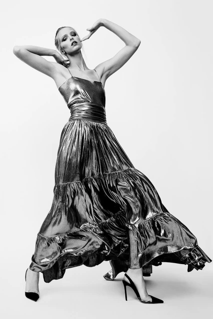 An adult female fashion model in a metallic long dress and high heels, striking a pose against a bright white backdrop in a photo studio.