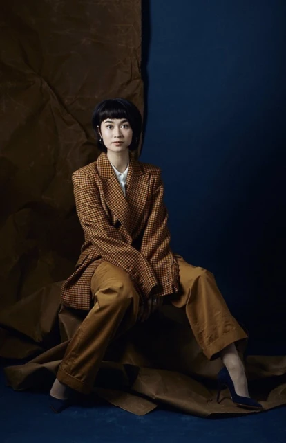 A stylish young woman with short dark hair poses in a brown checkered blazer and wide-leg trousers against a split blue and brown backdrop.