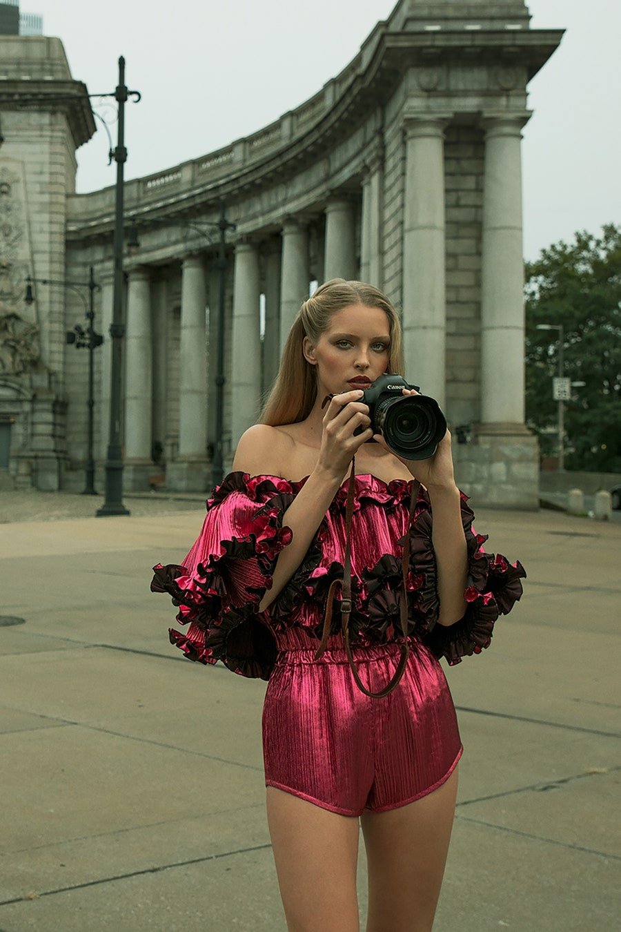 A woman in a red ruffled romper is photographed at eye level in an urban outdoor setting holding a professional digital camera.