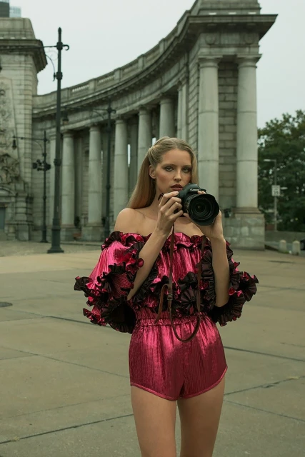 A woman in a red ruffled romper is photographed at eye level in an urban outdoor setting holding a professional digital camera.