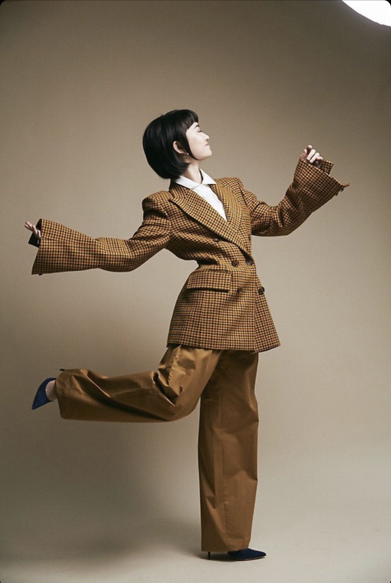 A woman with short black hair is wearing a tan plaid suit with wide-legged pants and blue shoes. She is striking a pose in a photo studio.