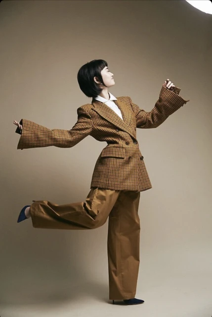 A woman with short black hair is wearing a tan plaid suit with wide-legged pants and blue shoes. She is striking a pose in a photo studio.