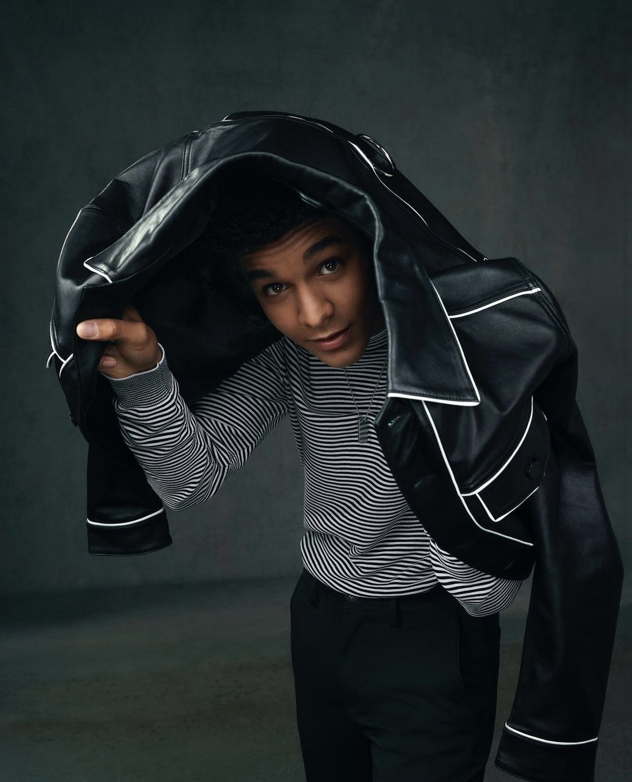 Portrait of a young adult male under a black leather jacket with white trim, wearing a striped sweater, against a grey backdrop.