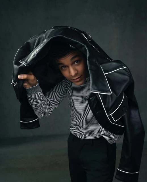 Portrait of a young adult male under a black leather jacket with white trim, wearing a striped sweater, against a grey backdrop.