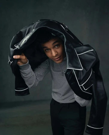 Portrait of a young adult male under a black leather jacket with white trim, wearing a striped sweater, against a grey backdrop.