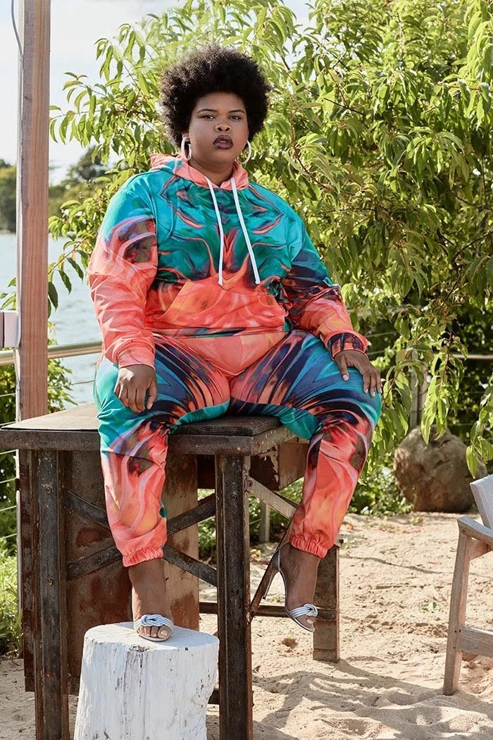 A woman with dark, voluminous hair poses outdoors wearing a colorfully patterned hoodie and sweatpants, seated on rustic furniture.