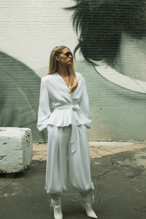 Model standing outdoors wearing a white silk blouse, white trousers, and white boots against a brick wall.