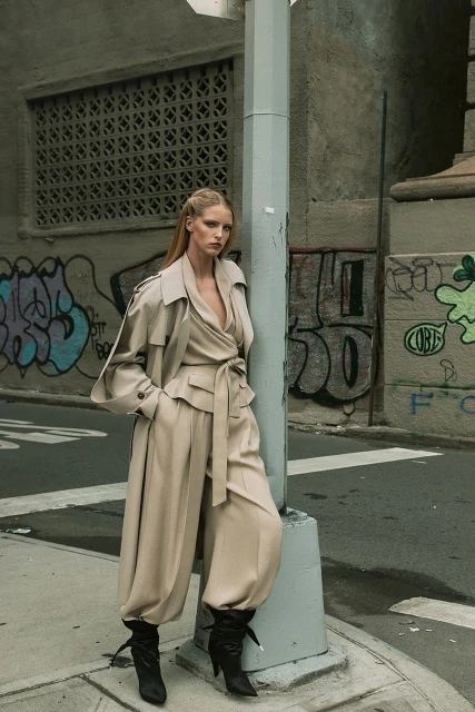Adult woman in a trench coat and baggy pants posing on a city street corner near a grey lamppost against a graffitied wall.