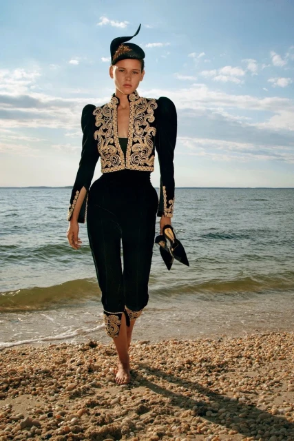 A fair-skinned woman walking barefoot on a beach, wearing a black velvet outfit with gold embroidery, holding black pointed toe heels.
