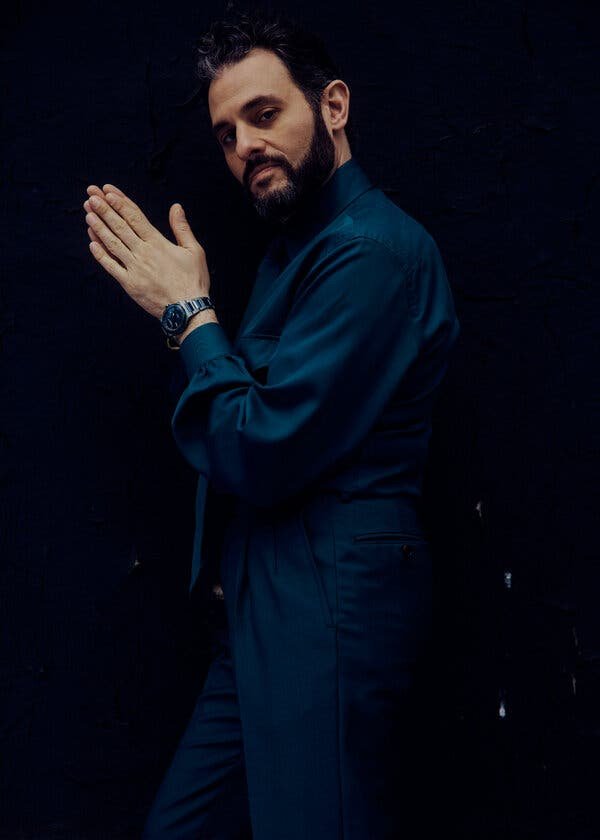 Studio shot of a bearded adult male in teal suit, posing with clasped hands against a dark backdrop, showcasing a watch.
