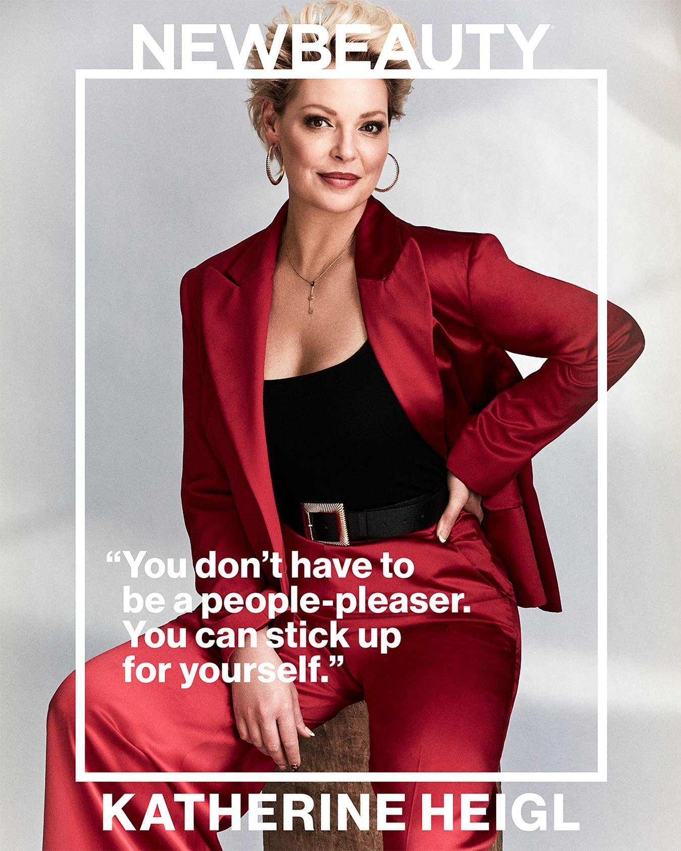 Katherine Heigl in a red satin suit poses for a portrait. She sits on a wooden stool in a photo studio setting, wearing gold hoop earrings.