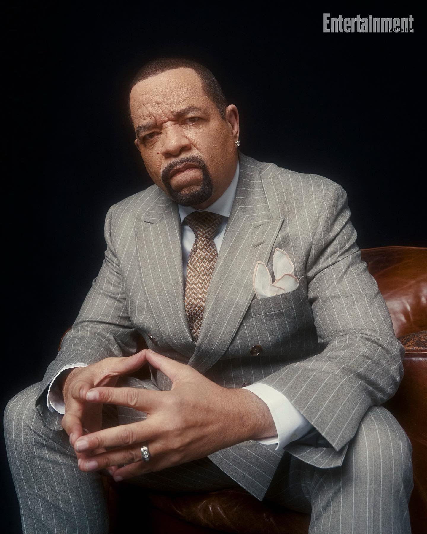 Portrait of Ice-T in a grey pinstripe suit and tie, seated in a brown leather chair against a dark backdrop, with Entertainment logo visible.