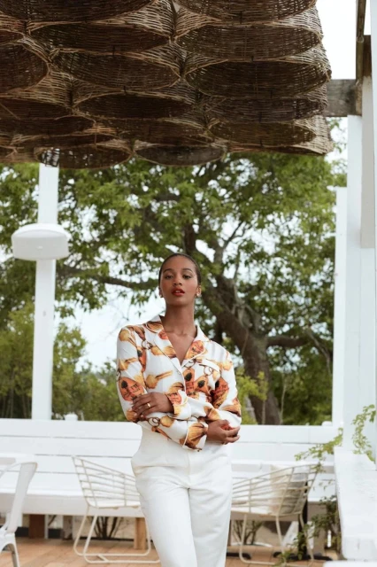 Elegant woman in a patterned blouse and white pants poses outdoors with a wicker canopy, white railings, and green foliage backdrop.
