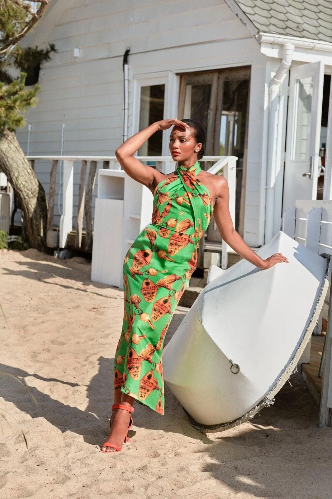 Elegant adult female model poses in a printed halter dress next to a boat on a sandy beach, with a white house in the background.