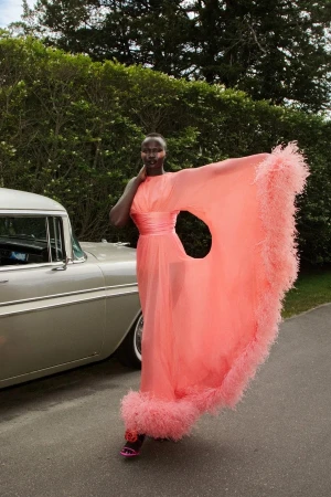 Black model wearing a flowing coral pink dress with feather trim, posing beside a vintage car and green trees.