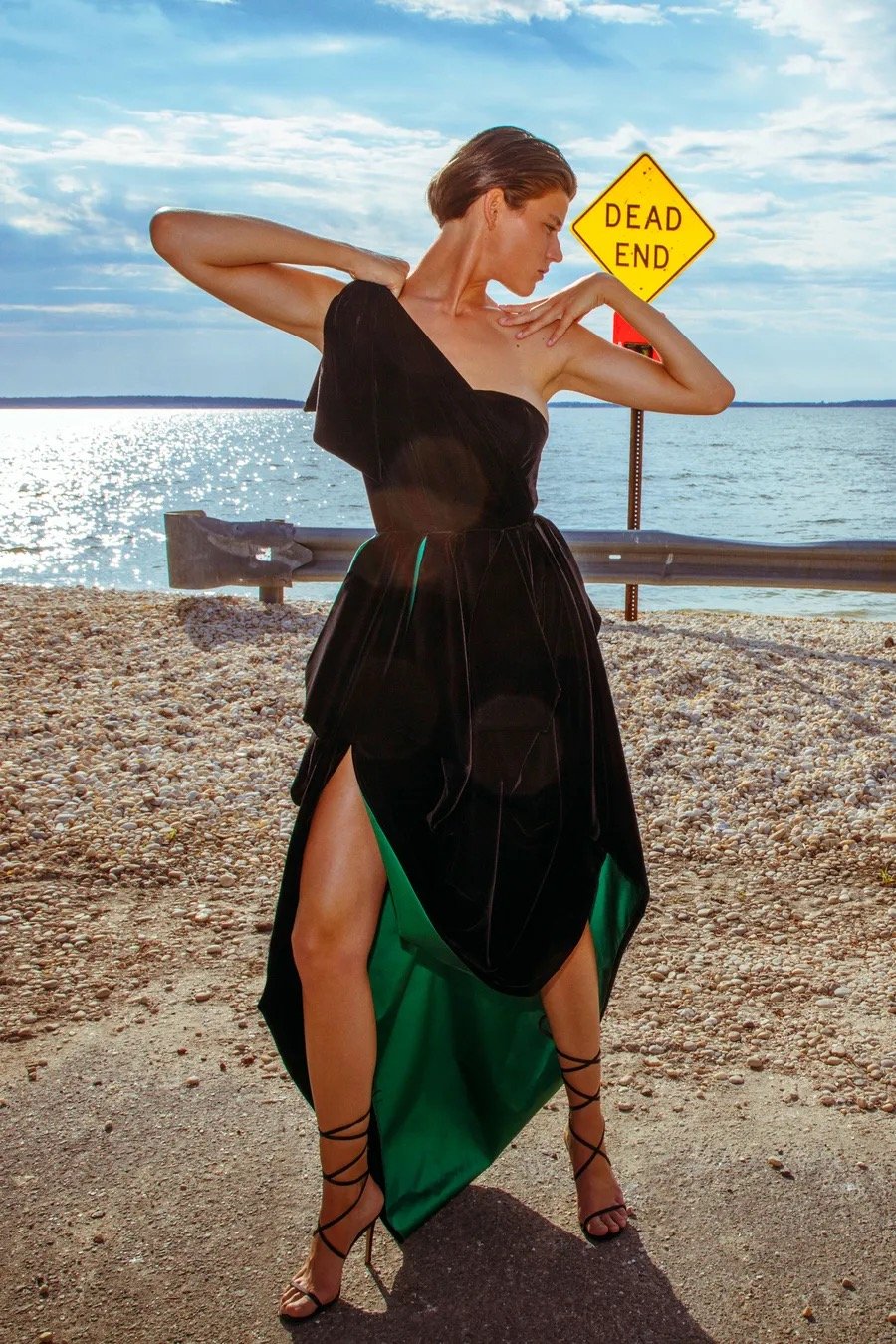 A fashionable woman in a black gown poses beside a yellow Dead End sign on an empty beach with a sea and bright sky background.