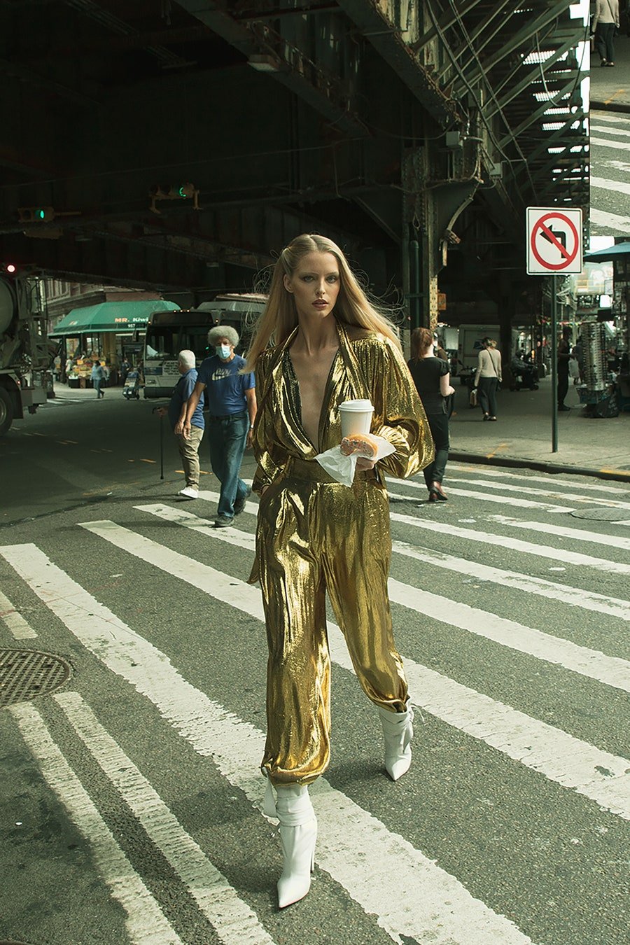 A woman in a shimmering gold jumpsuit walks across a city crosswalk, holding a coffee cup, with pedestrians and urban structures in the background.