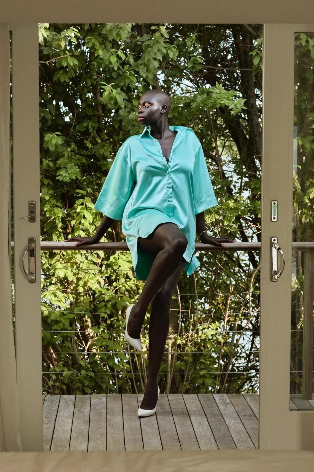 Elegant Black woman in teal oversized shirt and white heels poses on a deck with lush green trees in the background.