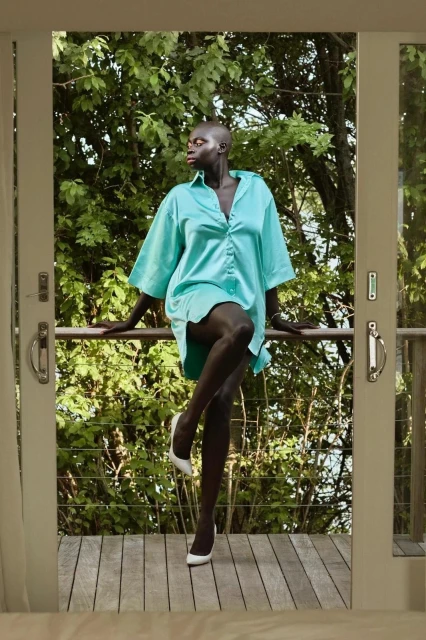 Elegant Black woman in teal oversized shirt and white heels poses on a deck with lush green trees in the background.