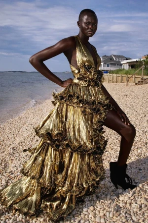 Model in a metallic gold pleated halter gown standing on a pebbled beach styled by Jared Depriest