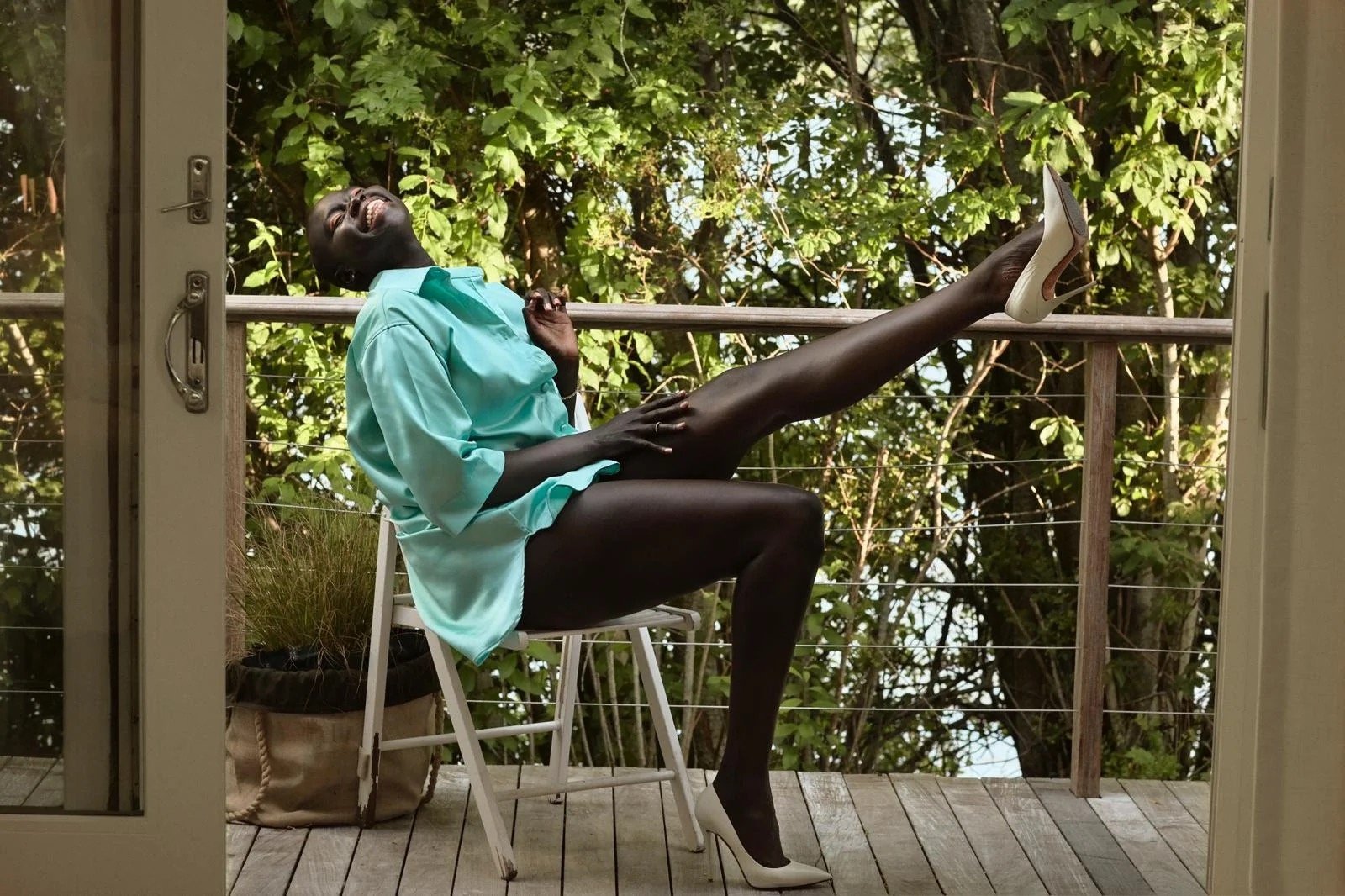 Dark-skinned woman in a turquoise shirt and white heels sits on a chair on a wooden deck with lush green trees in the background.
