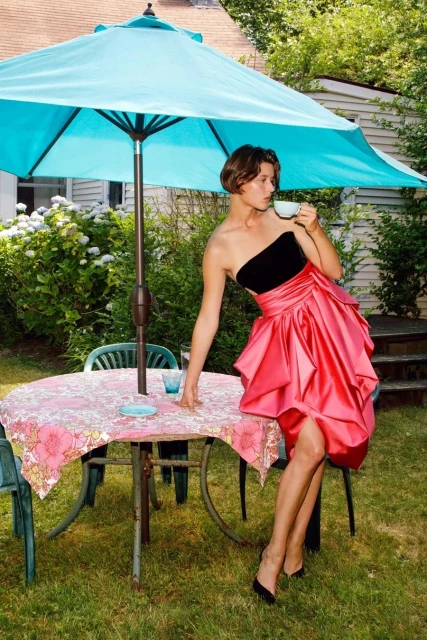 Young woman in a black strapless top and pink skirt sits on the edge of a table with a drink, under a teal umbrella in a green yard.