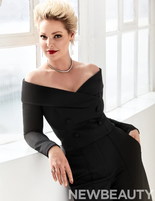Elegant woman in a black off-the-shoulder jumpsuit poses indoors by a window with bright, diffused natural light, wearing delicate jewelry.