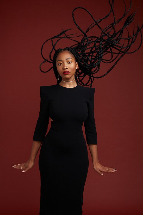 Portrait of a woman with long black braids wearing a black dress against a maroon background in a studio. Fashion shot.