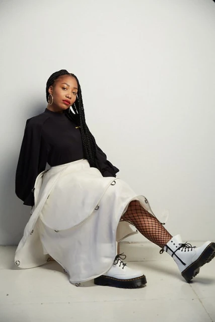 A young Black woman poses indoors with a black blouse, long braid, high-waisted white skirt, fishnet tights, and white combat boots in a studio.