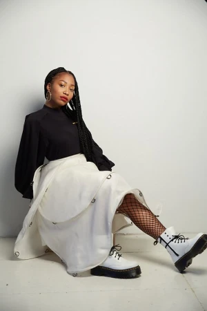 A young Black woman poses indoors with a black blouse, long braid, high-waisted white skirt, fishnet tights, and white combat boots in a studio.