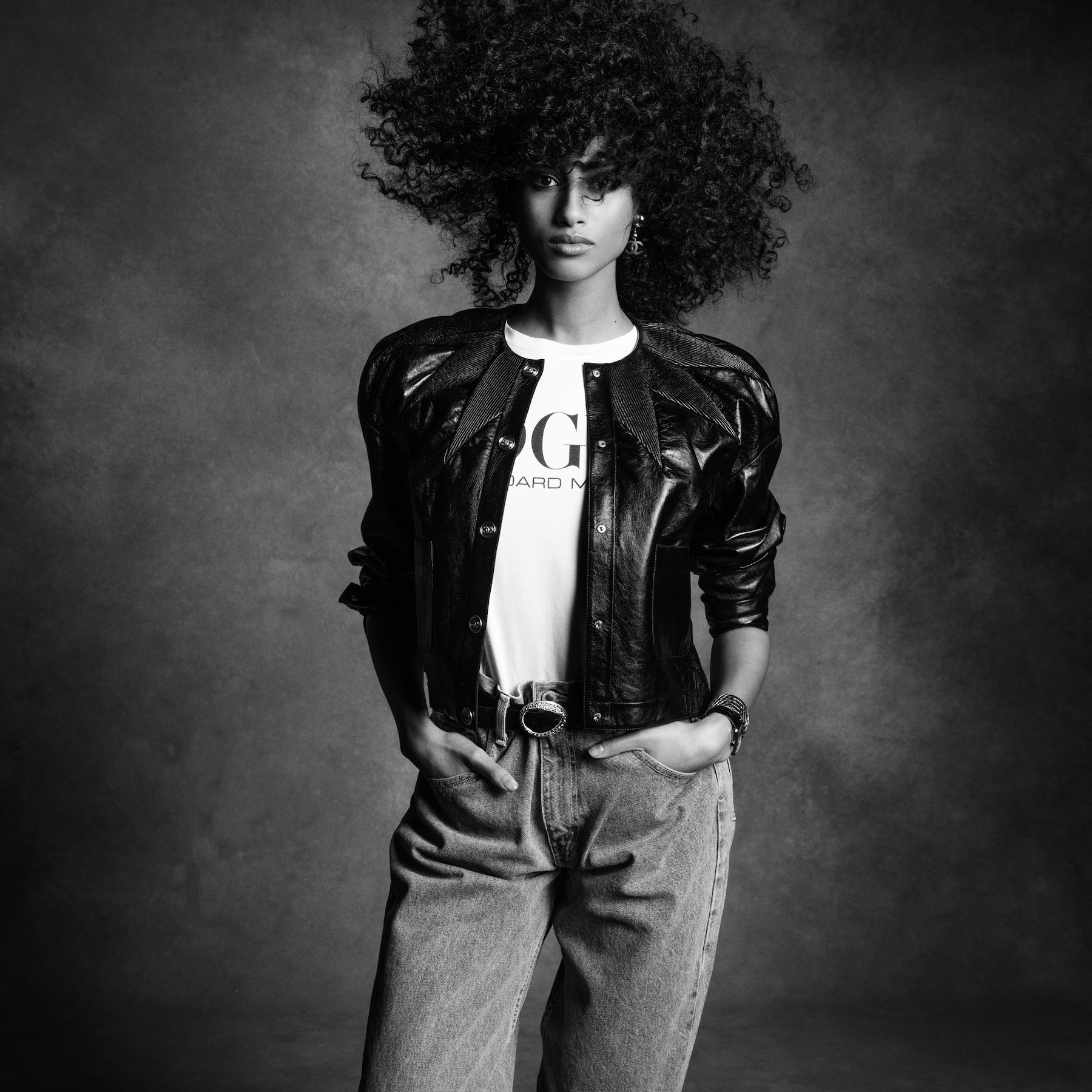Black and white portrait of a woman with afro wearing a black leather jacket, graphic tee, and light-wash denim jeans in studio.
