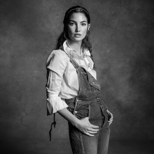 Black and white portrait of an adult woman wearing a white blouse and denim overalls, posed against a mottled dark background.