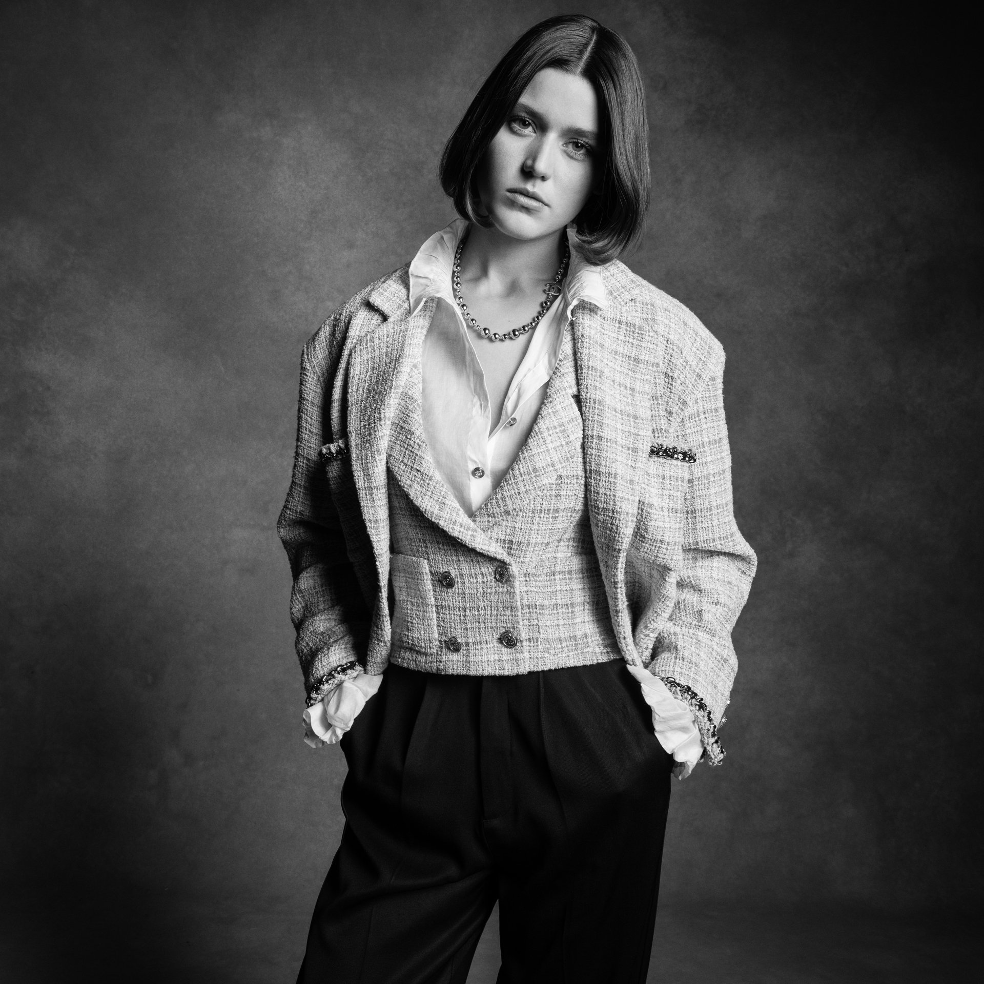 A woman in a tweed blazer and black pants poses against a textured gray background in a studio setting, showcasing a fashion-editorial look.