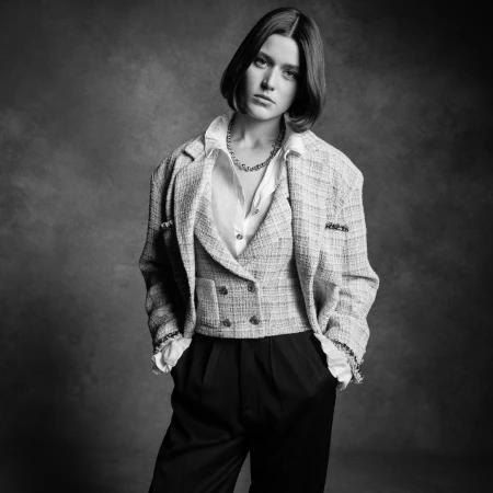 A woman in a tweed blazer and black pants poses against a textured gray background in a studio setting, showcasing a fashion-editorial look.
