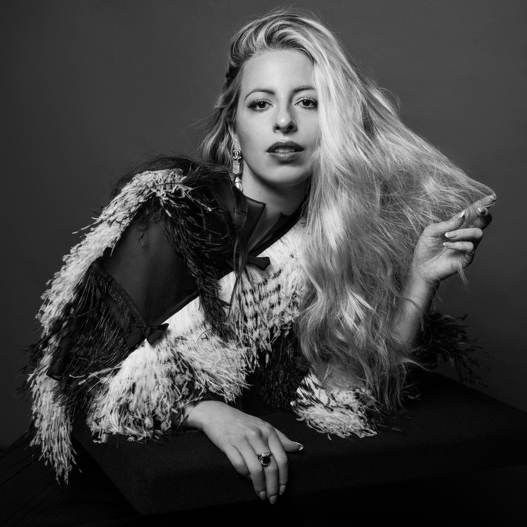 Black-and-white portrait of a blonde woman in a feathered jacket, looking at the camera with a neutral expression. Studio setting.