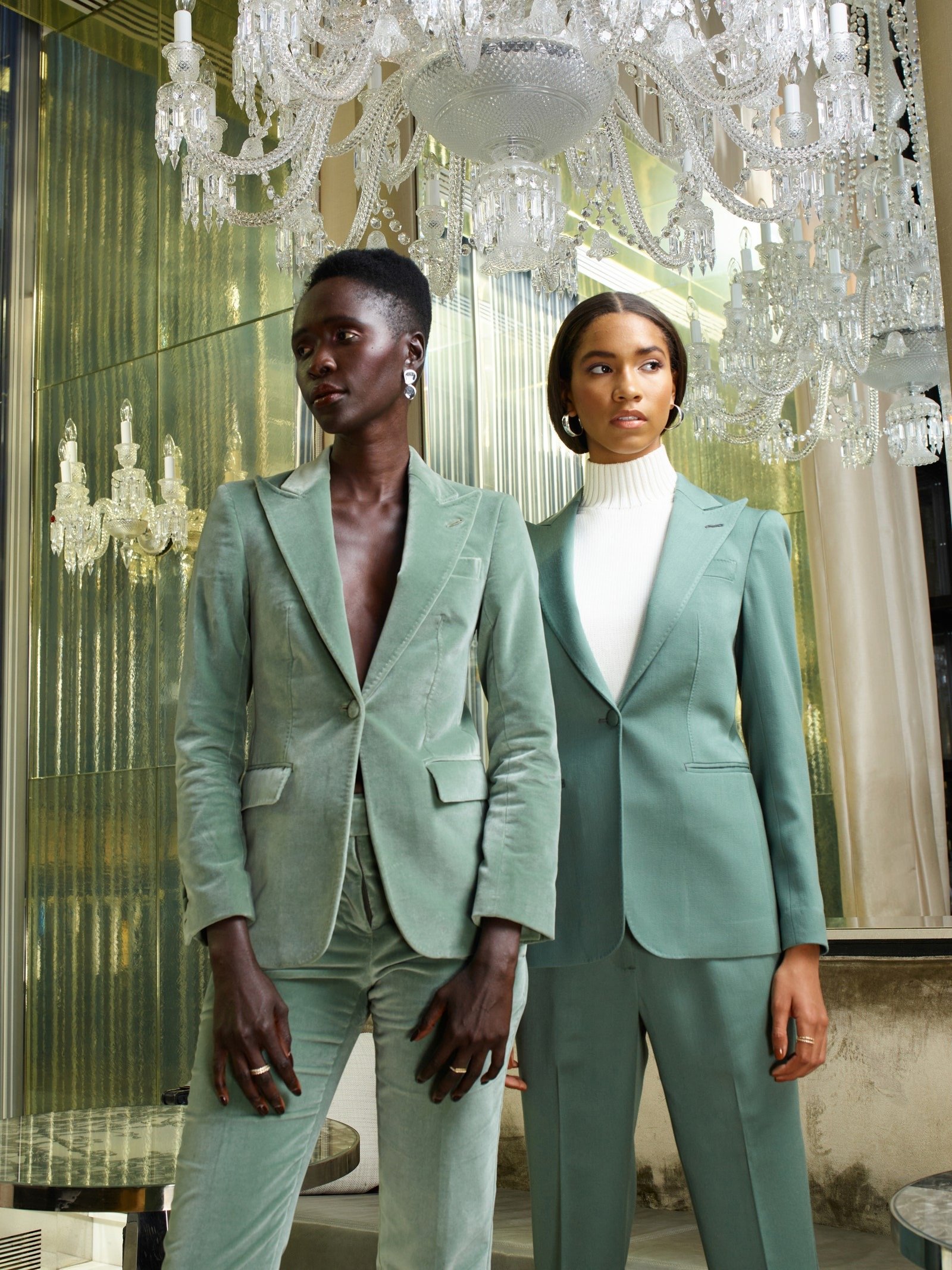 Two women in matching mint green pantsuits pose indoors under chandeliers in front of reflective wall panels for a fashion shot.