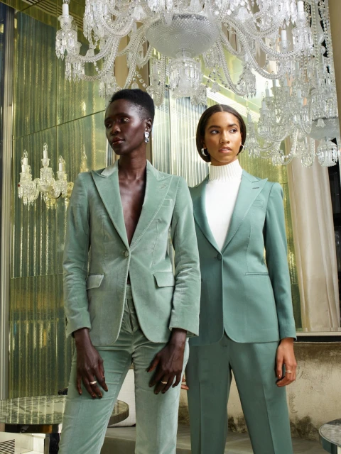 Two women in matching mint green pantsuits pose indoors under chandeliers in front of reflective wall panels for a fashion shot.