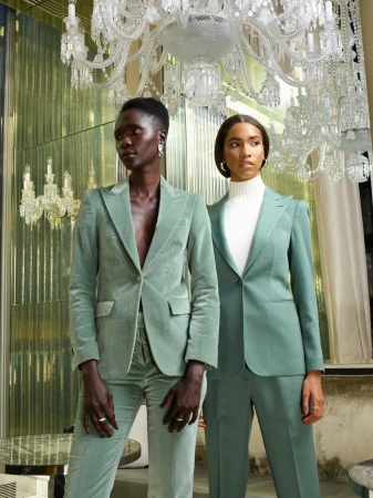 Two women in matching mint green pantsuits pose indoors under chandeliers in front of reflective wall panels for a fashion shot.