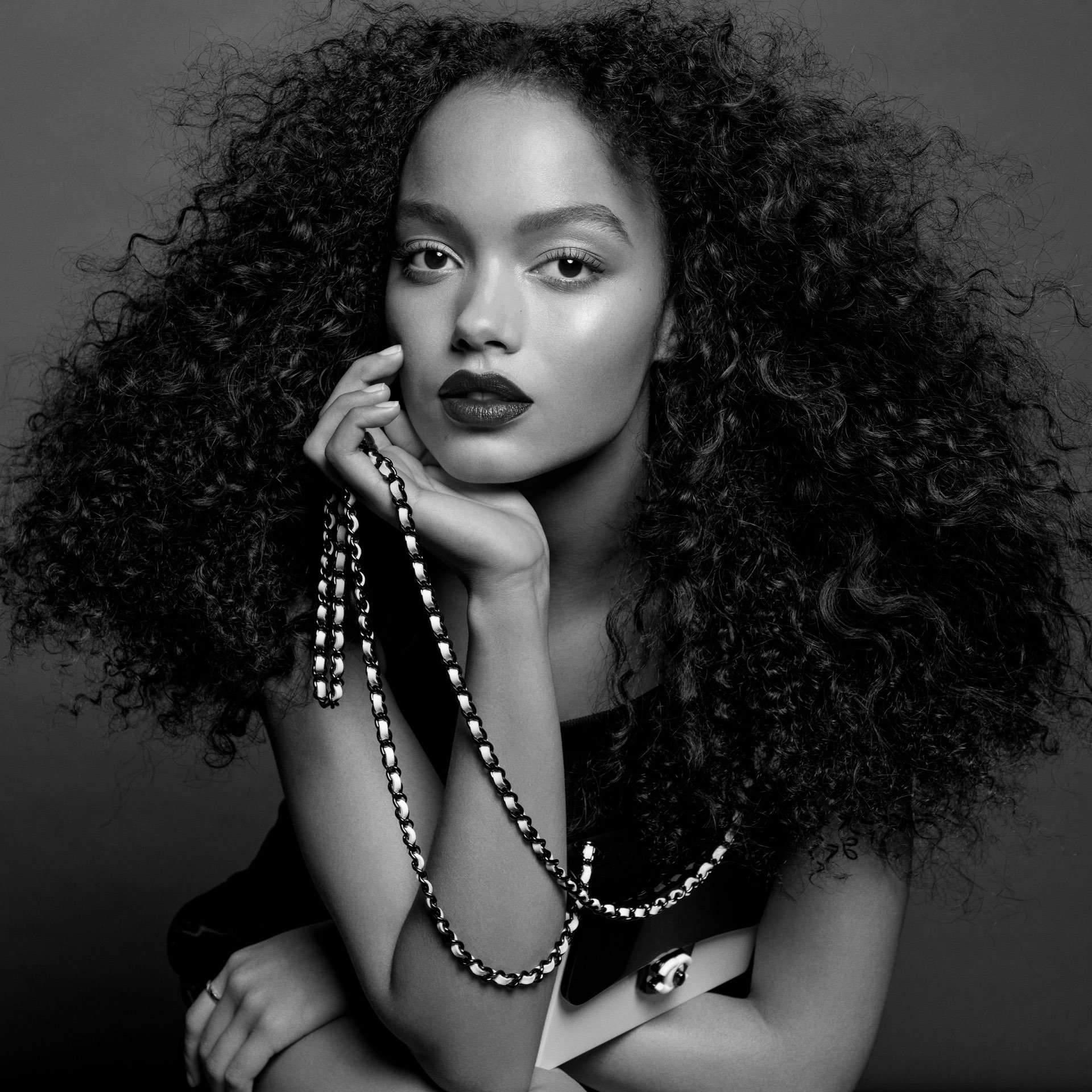 Black and white portrait of a woman with dark hair and lipstick holding a chain necklace against a neutral gray background.