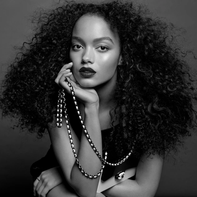 Black and white portrait of a woman with dark hair and lipstick holding a chain necklace against a neutral gray background.