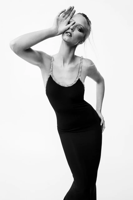 Black and white portrait of a woman wearing a black dress with embellished straps, hand near face, posing against a white backdrop.