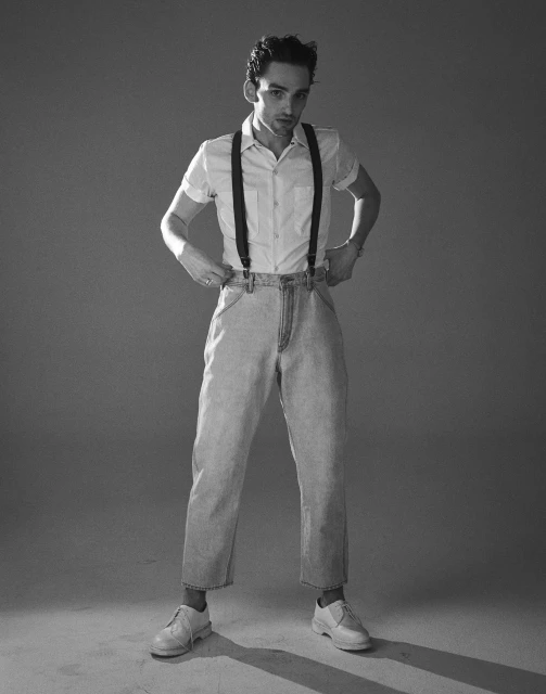 Black and white portrait of a young adult man in denim pants, white shirt, suspenders, and white shoes posing in a photo studio.