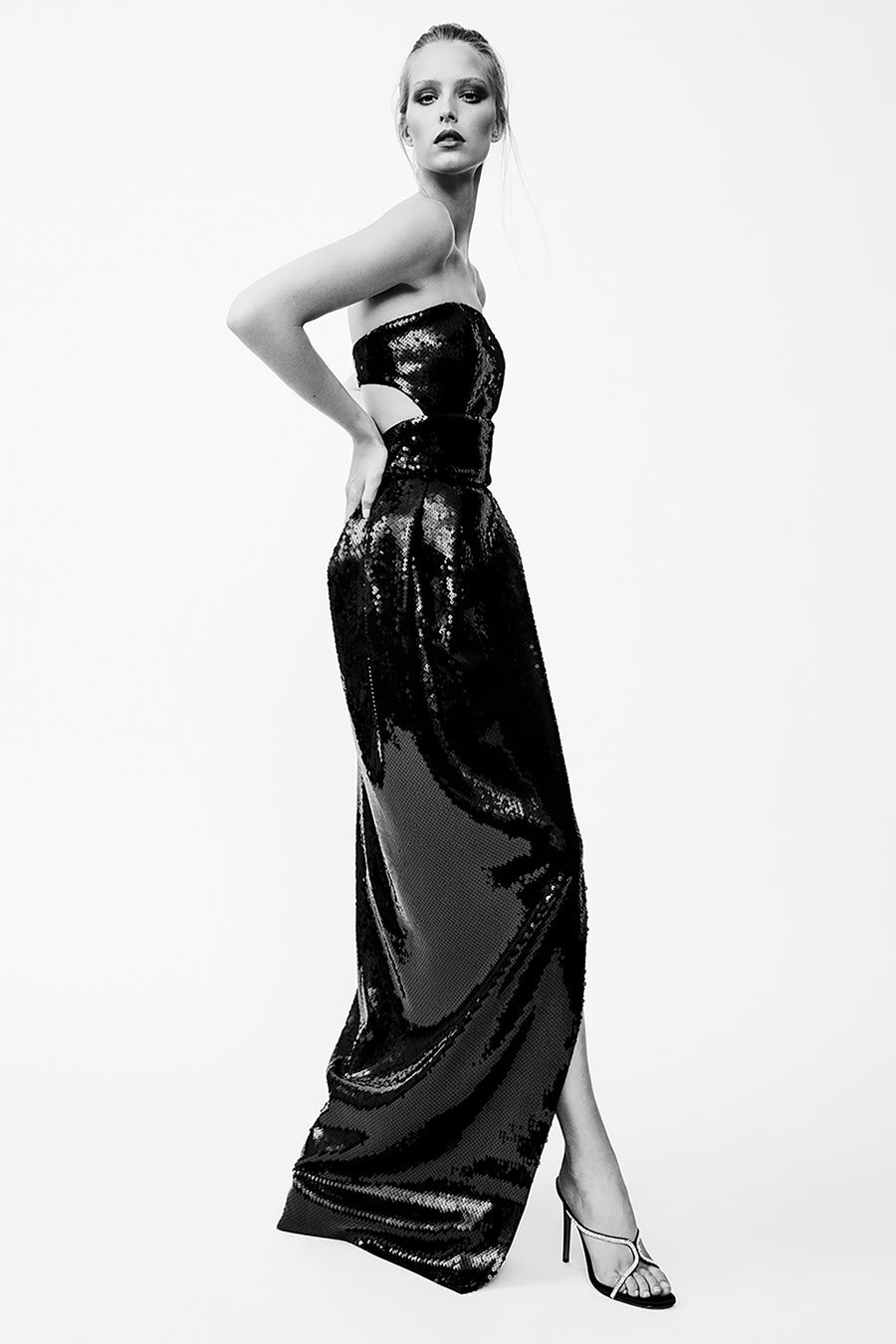 Black-and-white studio shot of an adult female fashion model posing in a sequined evening gown and high heels against a white background.
