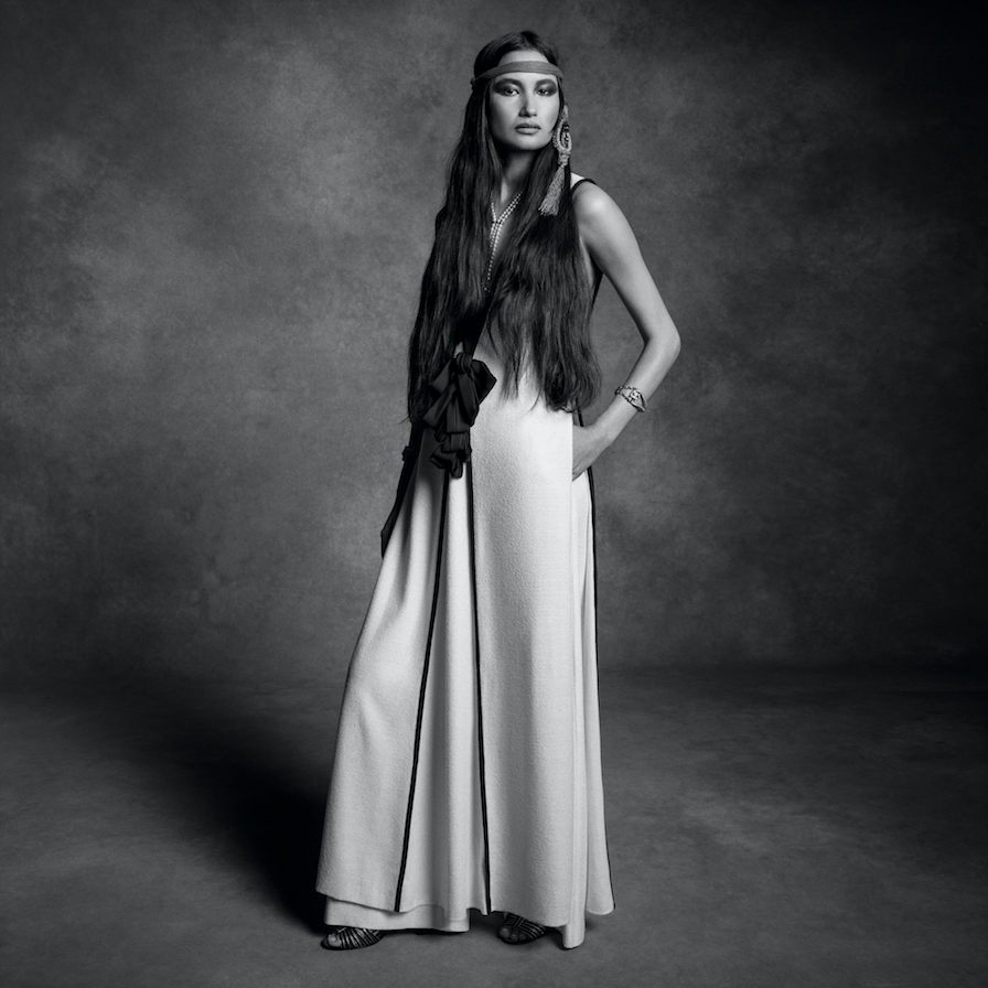 Model standing in a long gown and headband against a textured monochrome studio backdrop.