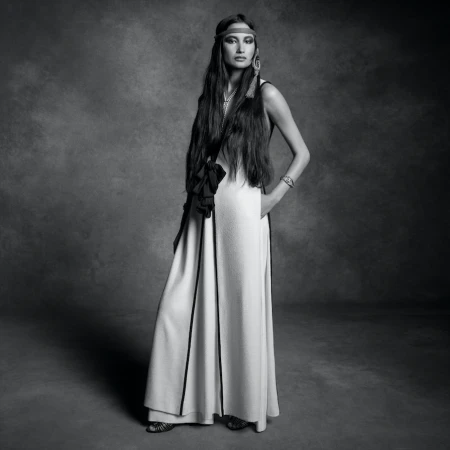 Model standing in a long gown and headband against a textured monochrome studio backdrop.