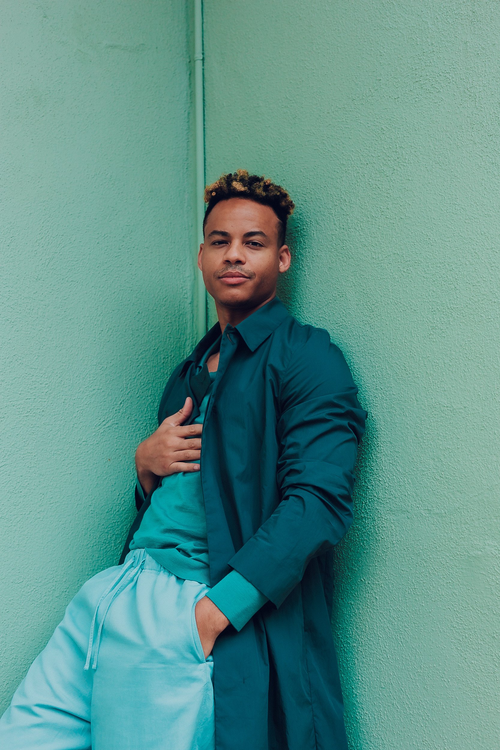 Young adult man with dyed hair poses against a green wall wearing a teal coat and light blue sweat suit. Casual fashion style.